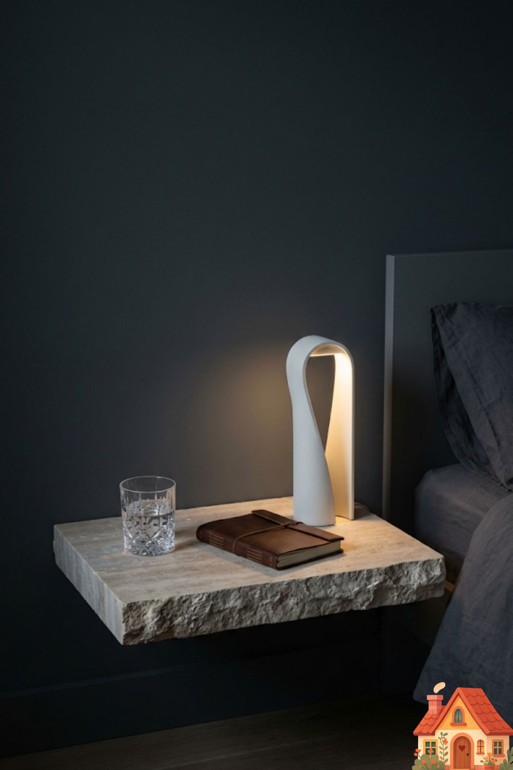 A minimalist bedroom setup. A thick, raw slab of honed travertine stone acts as a floating nightstand against a dark, moody wall. Resting on the stone is only a heavy crystal water glass, a leather-bound journal, and a sculptural, matte-white minimalist lamp. Absolute, curated perfection.